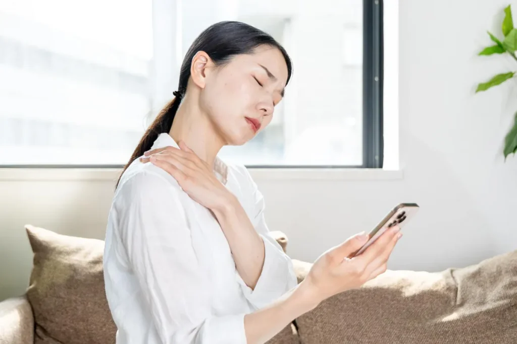 白いソファでスマホを見て首肩の痛みを感じる女性